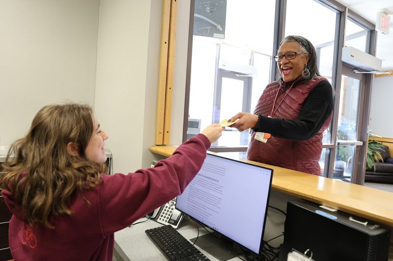 conference-services-summer A student working at the Housing Services front desk hands a document to another person who is standing on the opposite side of the front desk. A lobby with a couch and plants can be seen behind the guest. Both are wearing Hokie colors, a maroon sweatshirt and a maroon vest respectively, and smiling.