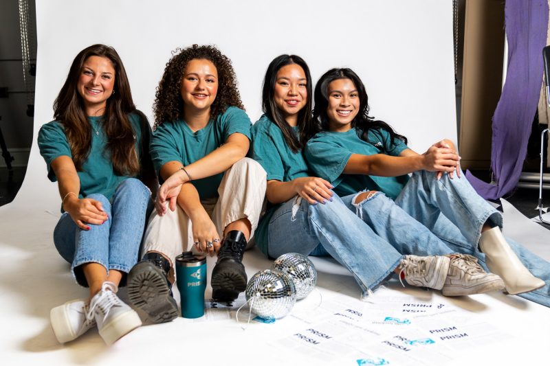 Four PRISM student leaders in teal shirts and jeans sit together in front of a white backdrop.