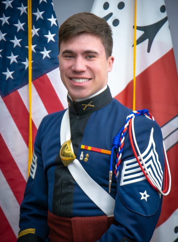 Portrait of Cadet Connor Christian in dress uniform in front of US and Corps flags.