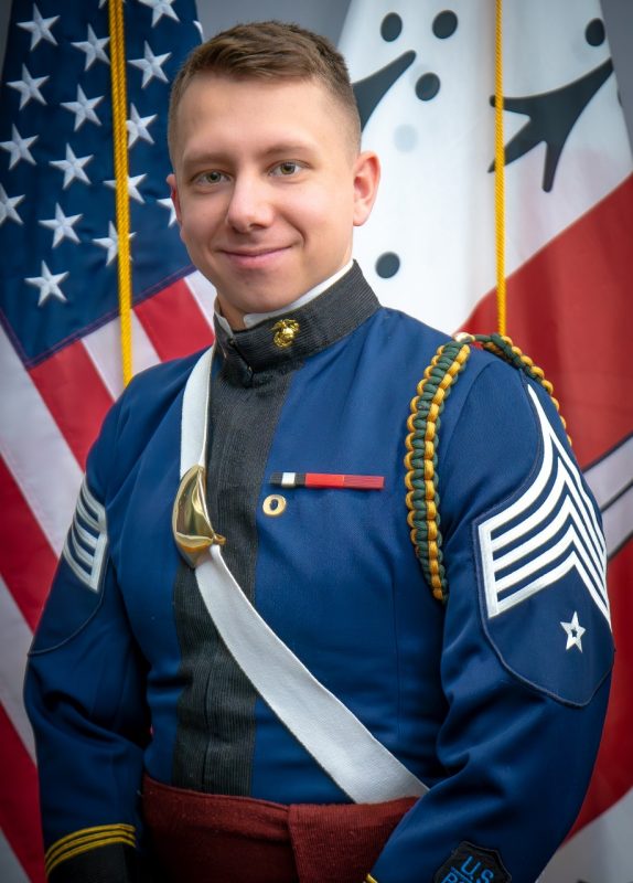 Portrait of Cadet Carson Barrette in dress uniform in front of US and Corps flags.