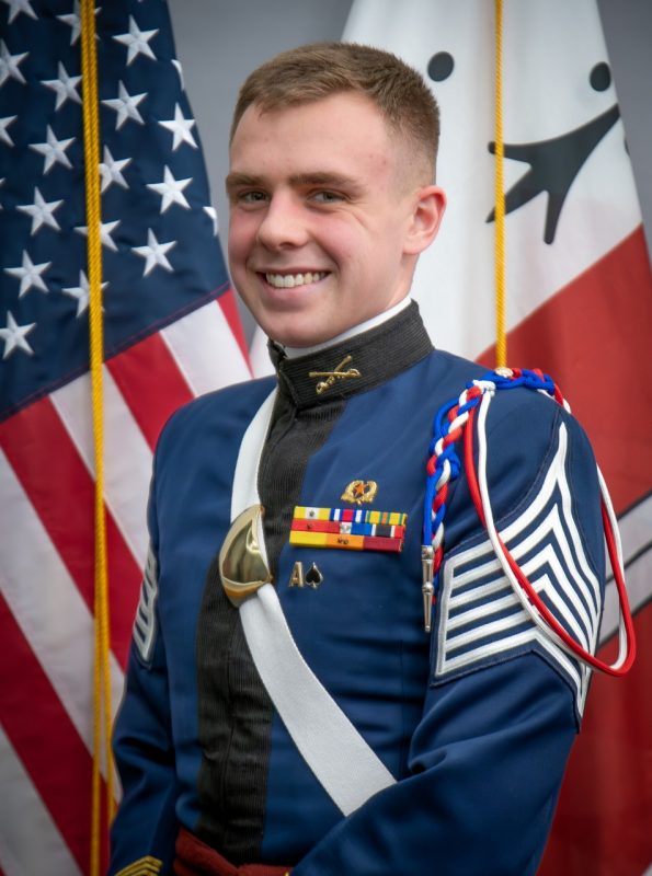 Portrait of Cadet Thomas Feely in dress uniform in front of US and Corps flags.