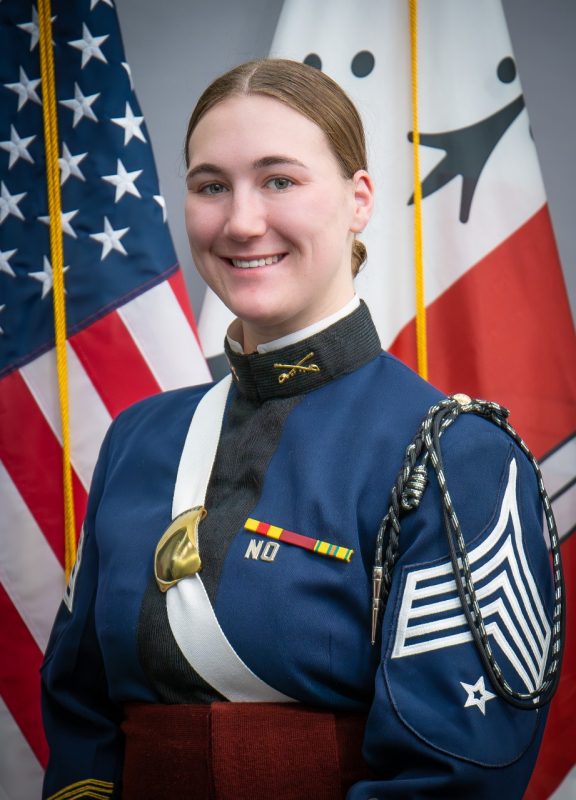 Portrait of Cadet Sidney Fredericks in dress uniform in front of US and Corps flags.