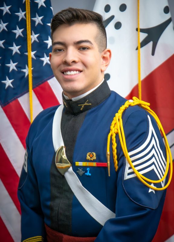 Portrait of CadetAiden Estrella in dress uniform in front of US and Corps flags.