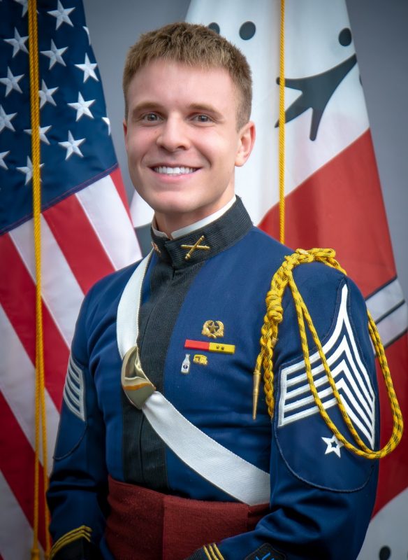 Portrait of Cadet Branden Gambone in dress uniform in front of US and Corps flags.