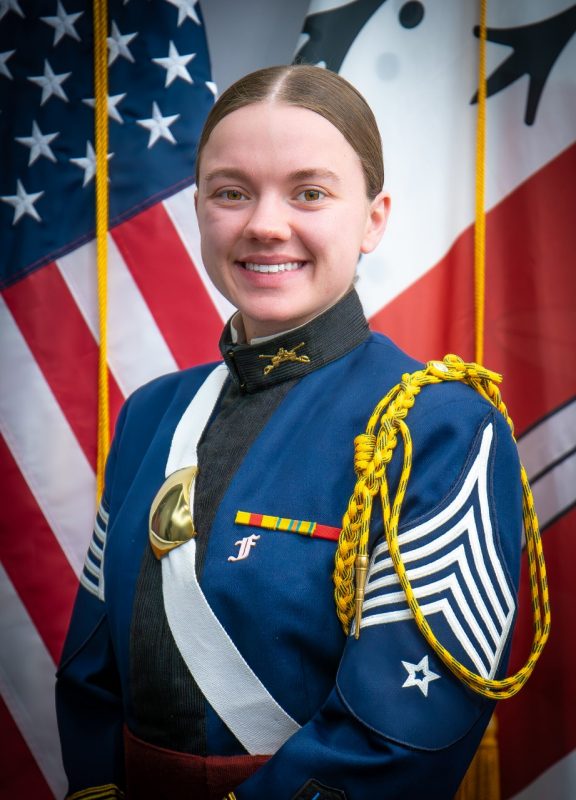 Portrait of Cadet Syerra Sylvester in dress uniform in front of US and Corps flags.