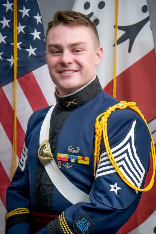 Hudson sits in his dress uniform in front of the American and Corps of Cadets flags.