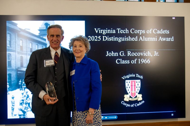 Rocovich and his wife stand together smiling in front of a screen that announces him as the award recipient. He is holding the shiny glass award.