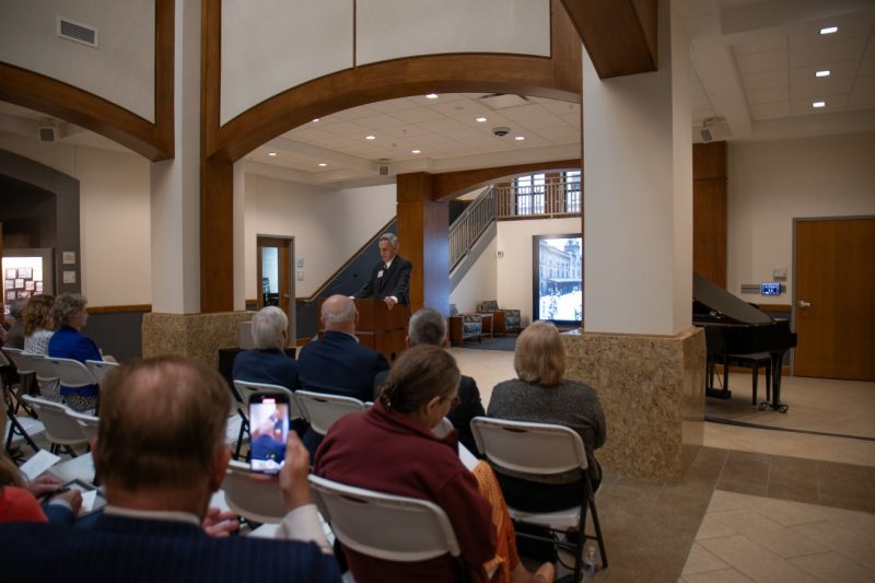 A crowd sits in an atrium with arched ceilings as Rocovich speaks at center from a podium