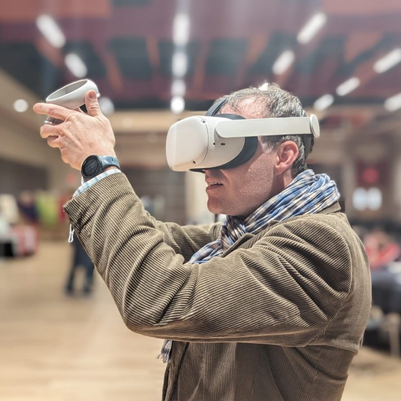 An International Mother Language Day virtual reality experience created by Ibrahim Tahmid. Photo courtesy of Ibrahim Tahmid. A man in a tan tweed jacket and a checkered blue and white scarf wears a white virtual reality headset. He holds a white contraption out in front of his face. There is a black watch with a blue strap on his wrist.