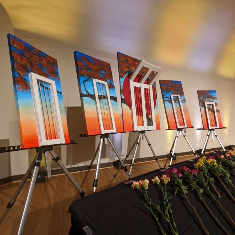 Five pieces from an art installation by Badhan Das at an International Mother Language Day event. Photo courtesy of Ibrahim Tahmid. In an open-concept art space with white walls and hardwood flooring, five canvases are positioned in front of a black table where pink, orange, yellow, and purple flowers are laid. The canvases each have a painting of part of a tree against a sunset, overlayed by a white window frame. The five canvases fit together to depict a tree top at sunset. There is a large red circle in the center canvas, along with the tree top.