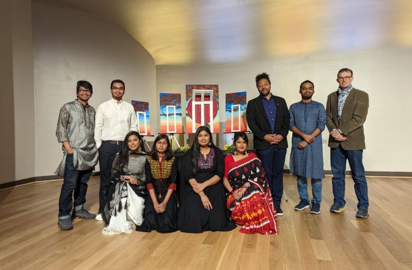 International Mother Language Day event organizers stand with Badhan Das's artwork. Photo courtesy of Ibrahim Tahmid. Nine individuals stand around an art installation in an open-concept art gallery with hardwood floors. The art installation features multiple canvases that fit together to depict a tree at sunset. Each of the canvases has a piece of the scene, as well as a white frame. The center canvas is the biggest, and features a large red circle in addition to the tree center and sunset background.