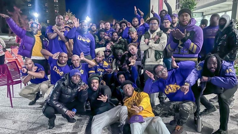 Members of the Omega Psi Phi Fraternity, Inc. with the Fall 2024 line of new members of the Eta Lambda chapter of the fraternity. Photo courtesy of Curtis Williams. Dozens of male students dressed in yellow and purple pose outside a building in the evening. They make silly faces and hand gestures, likely signifying their fraternity membership status. Behind them, the sky is dark blue, and two street lights shine brightly.