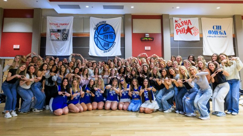 Alpha Delta Pi's 2025 new member class and chapter leaders at Bid Day in the Commonwealth Ballroom. Photo by Max Catalano for Virginia Tech. A group of more than 50 young women pose together in a gymnasium. Each of their outfits has at least one article of blue clothing, and their hands are forming a symbol of their sorority. Behind them, there are four banners for some of Virginia Tech's fraternity and sorority chapters.
