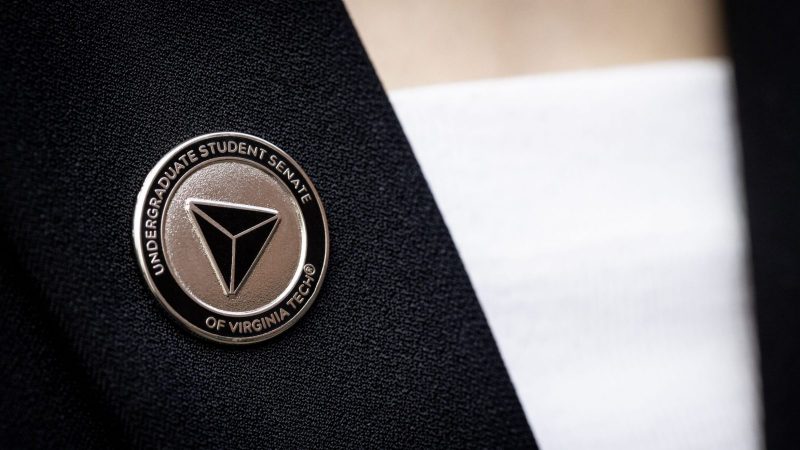 USS Pin A metal pin affixed to a black jacket over a white shirt. The pin is metallic with a black background and has a three-sided pyramid design in the center. "Undergraduate Student Senate of Virginia Tech" is engraved around the outside border of the pin.