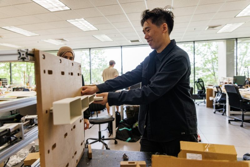 Sida Dai Designing Acoustic System A man works in a bright, modern classroom or workshop, adjusting white rectangular components attached to a vertical wooden board with various cutouts.