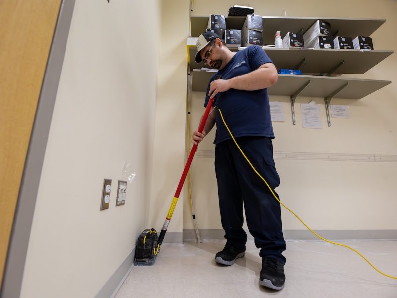 A custodian wearing a navy blue uniform and cap uses a floor cleaning machine to scrub along the edge of a wall in a supply room. Shelves stocked with boxes and cleaning supplies are mounted on the wall above them.