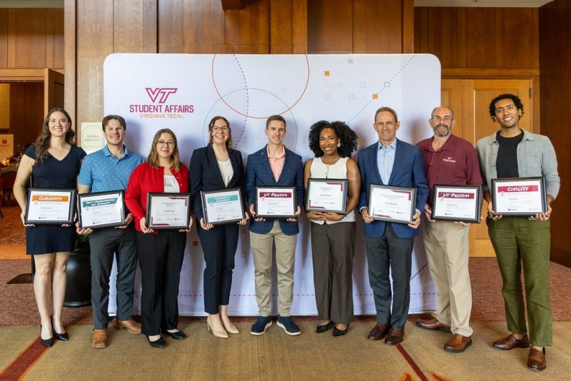 May Aspire! Award recipients and honorees. Photo by Brian Muncy for Virginia Tech. Nine smiling award recipients stand in front of a Virginia Tech Student Affairs-branded backdrop. They are all dressed in professional attire, and each holds their award certificate in front of them.