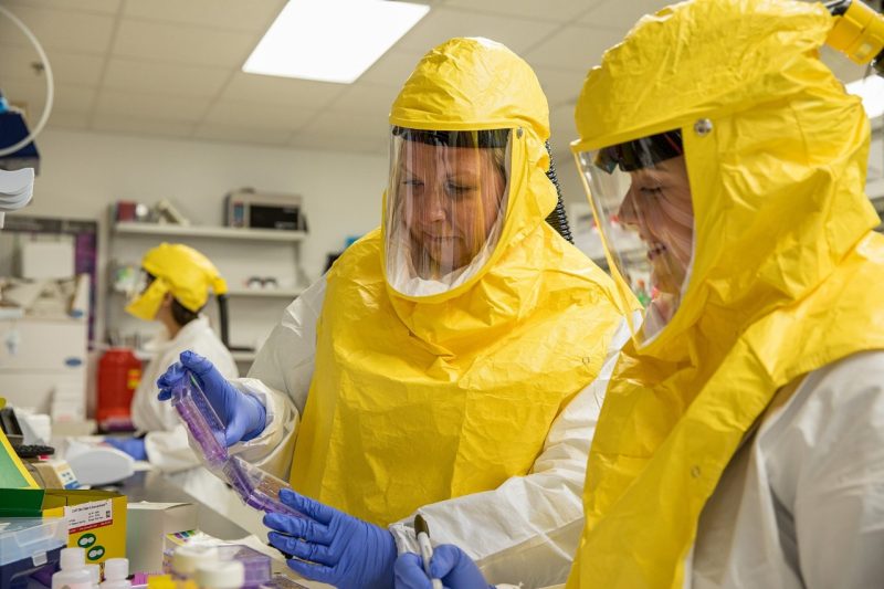 Faculty Awards Kylene Kehn-Hall (at center) in a laboratory setting teaching a student (at right) in personal protective equipment