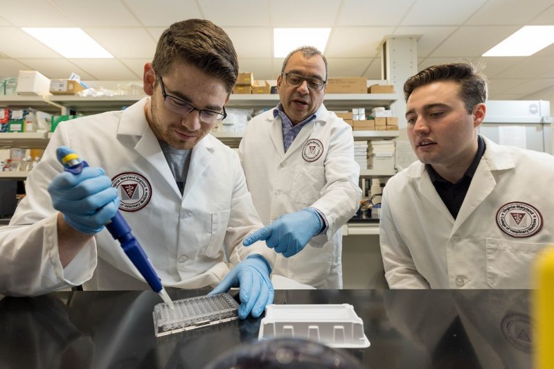 Faculty Awards (At center) Roger Ramirez-Barrios speaking with students (at left and right) in a laboratory setting. The student at left is using a pipette in the laboratory.