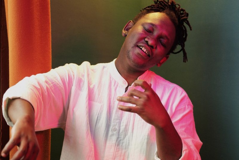 Abel Selaocoe, wearing a white shirt, performs with closed eyes and expressive hand gestures, bathed in warm pink and orange stage lighting.