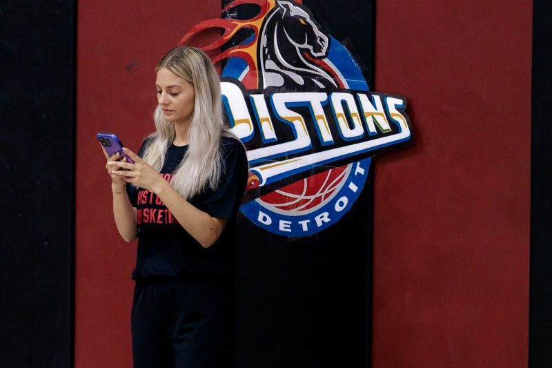 Emma Luis looks at her phone while standing in front of the Detroit Pistons logo on a wall.