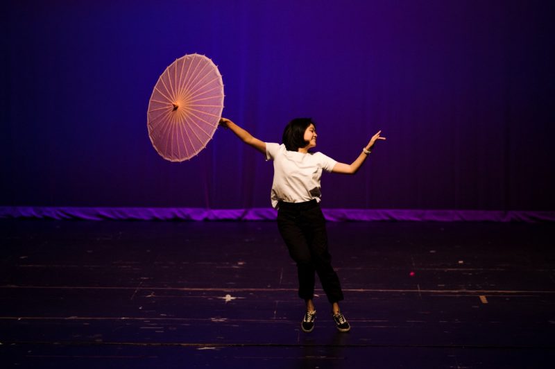 Asian Culture Show Fall 2022, student dancing on blue-backlit stage with a parasol. She is wearing a white shirt and black pants. 