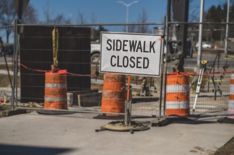 Signage reading "Sidewalk closed" in front of a gate and orange traffic cones. 