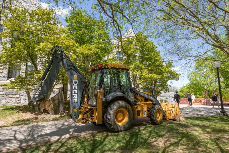 A yellow and black backhoe parked underneath green spring trees in front of a grey hokie stone building on a sunny day