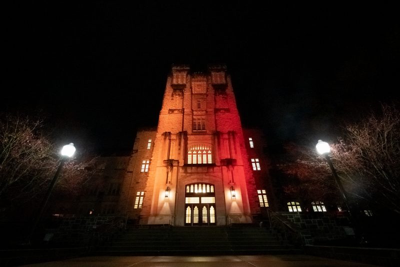 Grey Hokie Stone Burruss Hall illuminated orange on one side and maroon on the other side at nighttime.