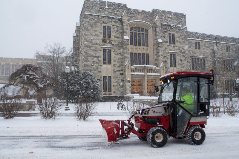 A tractor pushes snow out of a roadway in front of a grey Hokie Stone Building on a snowy day