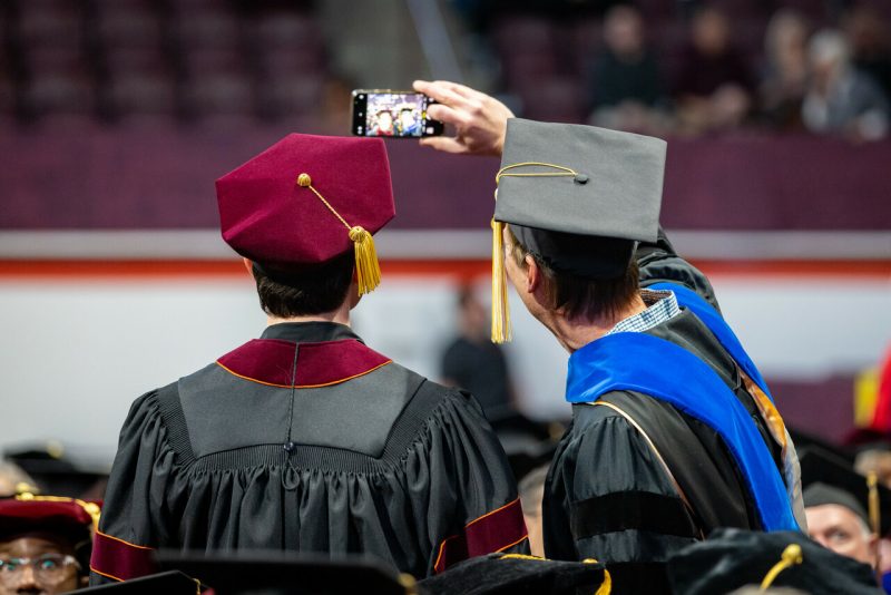 From behind, two students take a selfie wearing graduation regalia inside Cassell Coliseum