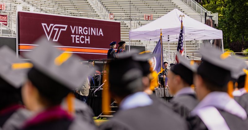 Many people wearing graduation caps and gowns sit in chairs watching people walk across an outdoor stage in a stadium. The Virginia Tech logo is on a sign in the background. 