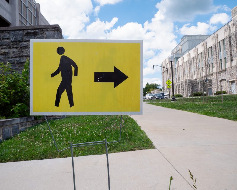 A yellow sign with a black pedestrian walking symbol and an arrow pointing right on it posted into the ground. A grey Hokie Stone building is in the background. 