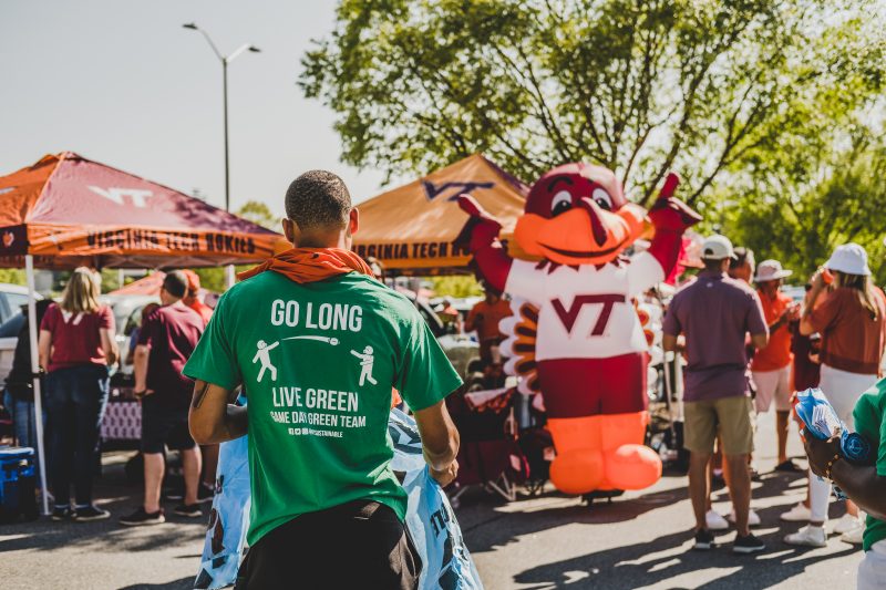 A person wearing a green t shirt that has writing on the back that says "Go long. Live green. Game day green team." carries many blue recycling plastic bags to a parking lot full of people tailgating with Virginia Tech paraphernalia. 