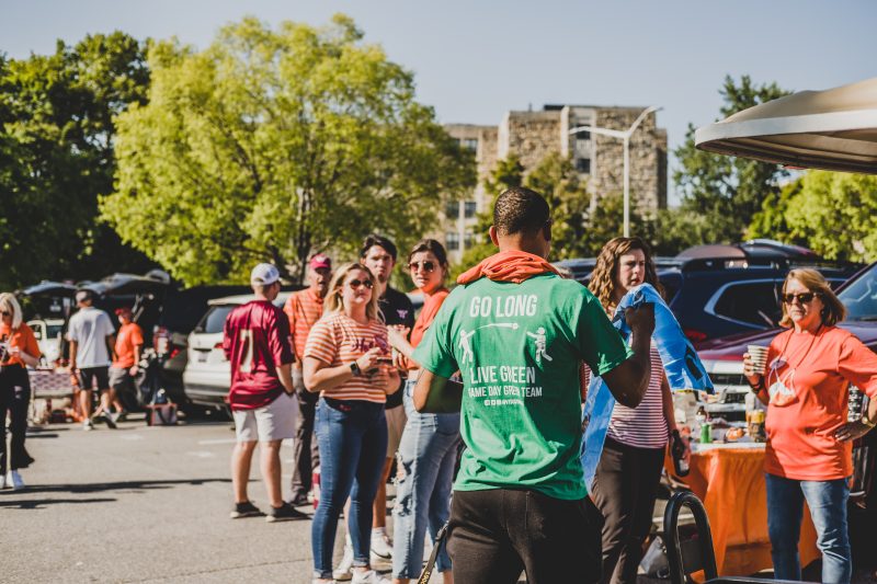 A person wearing a green t shirt that says go long live green passes out a blue recycling bag to tailgaters wearing orange and maroon in a parking lot