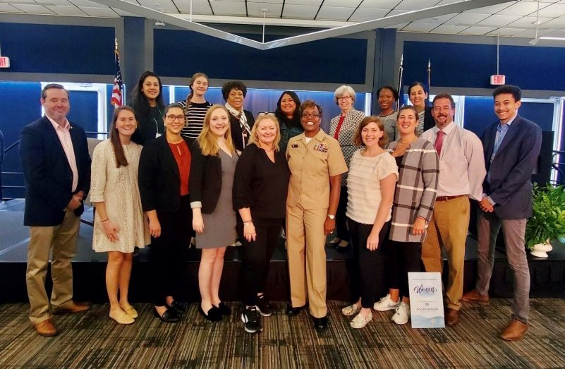 A group of people arranged in three rows smile at the camera.  Captain Janet Days stands in the center in a tan military uniform.