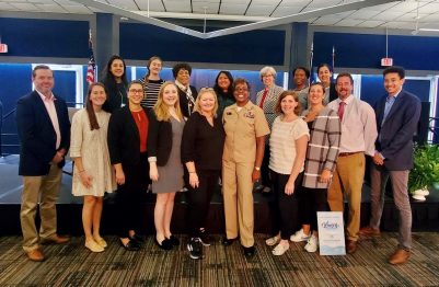 A group of people arranged in three rows smile at the camera.  Captain Janet Days stands in the center in a tan military uniform.