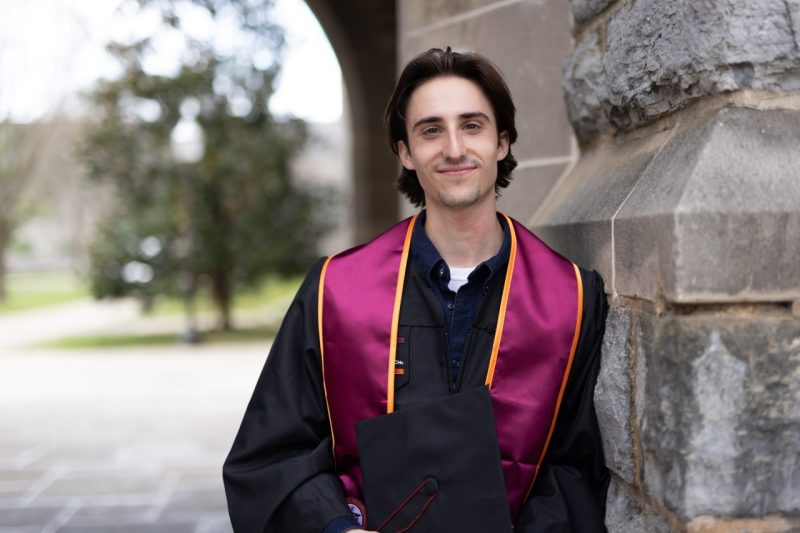 Wearing his graduation robe and stole, Jack smiles and leans against a Hokie Stone wall.